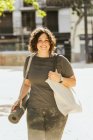 Positive young plus size female with curly dark hair in casual clothes smiling while walking on city street with rolled mat before yoga session in sunny morning — Stock Photo
