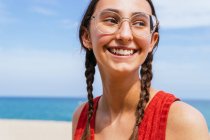 Mujer alegre en ropa de verano con coletas de pie en la orilla arenosa con el mar azul tranquilo en el día soleado - foto de stock