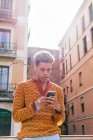 Young Hispanic guy with Afro hair in stylish colorful outfit browsing mobile phone while standing on railing near urban building in sunlight — Stock Photo