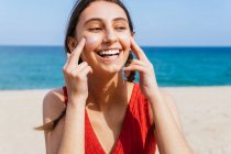Улыбающаяся женщина наносит лосьон для загара на лицо в солнечный день летом на пляже — стоковое фото