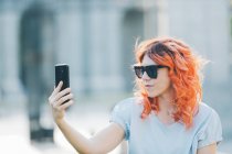 Модная женщина с рыжими волосами и в солнечных очках, снимающая себя на мобильном телефоне в солнечный день на улице — стоковое фото