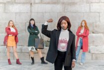 Уверенная афроамериканка показывает бицепс, стоя против группы многорасовых женщин, демонстрирующих концепцию женской силы — стоковое фото