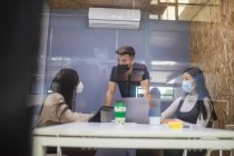 A través de un vaso de compañía de compañeros de trabajo multirraciales en máscaras protectoras reuniéndose en la mesa y discutiendo proyecto durante reunión de negocios - foto de stock