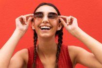 Encantada joven hembra con coletas que se pone gafas de sol de moda contra el fondo rojo en la calle de la ciudad - foto de stock