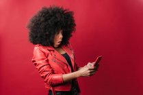 Emocionada mujer afroamericana con peinado afro que navega por el teléfono móvil sobre fondo rojo en el estudio - foto de stock