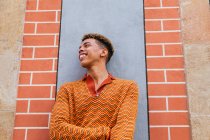 Young stylish cheerful ethnic curly haired guy in trendy outfit leaning against wall on urban street looking away — Stock Photo