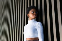 Unemotional African American female in trendy sweater looking away against striped building wall on street — Stock Photo