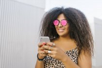 Mulher jovem e afro com óculos de sol conversando com seu telefone inteligente e sorriso — Fotografia de Stock