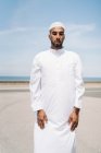 Voller Junge islamischer Mann in traditioneller weißer Kleidung steht auf Teppich und betet gegen den blauen Himmel am Strand — Stockfoto