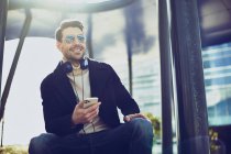 Вміст чоловіка в стильному одязі з навушниками та мобільними телефонами, що сидять у місті в сонячний день — стокове фото