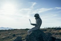 Homem astronauta de corpo inteiro em trajes espaciais navegando dados no netbook enquanto sentado na rocha fora da estação com antenas em forma de foguete — Fotografia de Stock