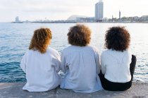 Rückansicht anonymer Freunde mit lockigem Haar, die dicht an Stadtbild und Böschung im Sonnenlicht sitzen — Stockfoto