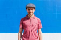 Радостный молодой бородатый мужчина в стильной повседневной одежде и кепке наслаждается летним днем возле синей стены на городской улице, глядя в камеру — стоковое фото