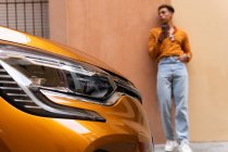 Junger stylischer ethnischer Lockenkopf im trendigen Outfit mit Smartphone, während er sich in der Nähe eines geparkten modernen orangefarbenen Autos an die Wand lehnt — Stockfoto