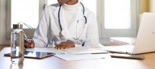 Обрізати невизначену чорну жінку-лікарку зі стетоскопом, пишучи інформацію на паперовому аркуші, готуючи медичний звіт за столом в офісі сучасної клініки — стокове фото