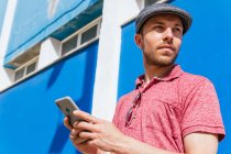 Знизу вдумливий молодий бородатий хіпстер у повсякденній сорочці поло та кепці переглядає мобільний телефон, стоячи на синій стіні на сонячному світлі — стокове фото