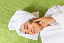 Top view happy young female with towel on head smiling and massaging face with jade roller during skin care routine at home — Stock Photo