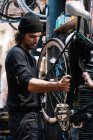 Vista lateral del joven maestro masculino examinando neumático en rueda de bicicleta mientras trabaja en taller de servicio de reparación profesional - foto de stock