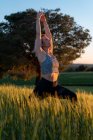 Adult female in sportswear practicing yoga with raised arms while looking up in countryside field — Stock Photo