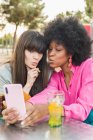 Багаторасова пара гомосексуальних жінок знімає себе на мобільний телефон і виливає губи, сидячи за столом з коктейлем у вуличному кафе — стокове фото
