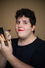 Женский андрогинный мужчина с вьющимися волосами наносит помаду на губы, делая макияж — стоковое фото
