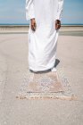 Cultiver mâle islamique en vêtements blancs traditionnels debout sur le tapis et prier contre le ciel bleu sur la plage — Photo de stock