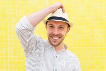 Feliz joven barbudo hipster chico en traje de moda y sombrero sonriendo mirando a la cámara mientras se enfría solo contra la pared amarilla en el día de verano - foto de stock