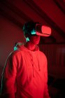 Человек в капюшоне во время использования VR очков и стоя в студии с красными неоновыми огнями — стоковое фото