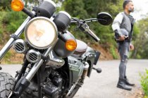 Серйозний чоловічий велосипедист у джинсах та шкіряній куртці тримає шолом у руці, стоячи на асфальтній дорозі біля припаркованого сучасного мотоцикла — стокове фото