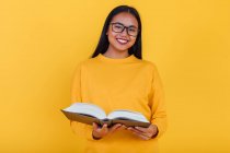 Inteligente alegre asiática estudante em óculos lendo livro didático e se preparando para o exame no fundo amarelo no estúdio olhando para a câmera — Fotografia de Stock