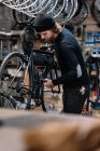 Vista lateral do jovem mecânico masculino qualificado com roda de cadeia de montagem de ferramentas na bicicleta durante trabalhos de reparo em oficina — Fotografia de Stock