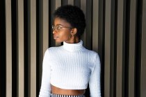 Unbewegte Afroamerikanerin im trendigen Pullover blickt gegen gestreifte Hauswand auf der Straße — Stockfoto