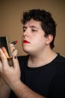 Homme androgyne féminin avec des cheveux bouclés appliquant du rouge à lèvres sur les lèvres tout en faisant du maquillage — Photo de stock