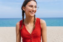 Cheerful female in summer clothes with pigtails standing on sandy shore with calm blue sea on sunny day — Stock Photo