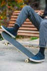 Homem irreconhecível em tênis equilibrando no skate enquanto sentado no banco de madeira no parque com arbustos durante o treinamento na cidade — Fotografia de Stock