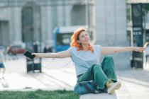Dreamy female with red hair sitting in street and listening to music in earphones while enjoying songs with closed eyes and outstretched arms — Stock Photo