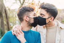 Amar casal LGBT de homens usando máscaras protetoras abraçando no parque durante a epidemia de coronavírus e beijando — Fotografia de Stock