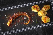 Top view of fried octopus tentacle and pieces of potato served with spices on black board on table — Stock Photo