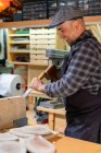 Боковой вид на веселого зрелого мастера с помощью молотка и зубила при создании деревянных деталей в столярной мастерской — стоковое фото