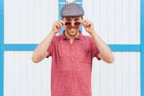 Young bearded guy wearing casual red polo shirt and cap looking at camera over sunglasses and smiling friendly while standing against wall on street in summer day — Stock Photo