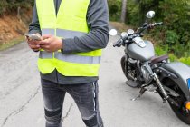 Crop ciclista masculino en chaleco de mensajería en el teléfono móvil, mientras que de pie cerca de la motocicleta rota cerca de exuberantes bosques verdes - foto de stock