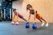 Young determined female athletes in sportswear standing in plank pose during functional training with weights on floor in gym — Stock Photo