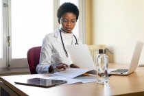 Jovem médica afro-americana em roupão branco com estetoscópio sentado à mesa com laptop e lendo registros médicos enquanto trabalhava no consultório da clínica — Fotografia de Stock