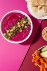 Draufsicht auf appetitliche Rote-Bete-Hummus garniert mit Kichererbsen auf zwei farbigen Hintergrund mit Brot und frischen Möhren-Gurken-Sticks serviert — Stockfoto