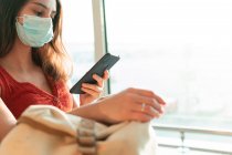 Женщина-туристка в защитной маске сидит в зале вылета аэропорта и ждет полета во время эпидемии коронавируса при использовании смартфона — стоковое фото