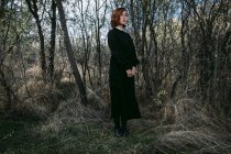 Жінка без емоцій у довгій чорній сукні, що стоїть в осінньому лісі під час святкування Хеллоуїна — стокове фото