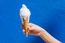 Mano de corte de hembra anónima con helado derretido en cono de gofre en día soleado sobre fondo azul - foto de stock