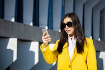 Mulher de negócios asiática com casaco amarelo e telefone inteligente andando na rua com edifício no fundo — Fotografia de Stock
