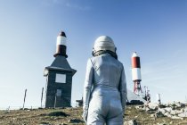 Voltar ver o homem no traje espacial em pé em solo rochoso contra cerca de metal e foguete listrado em forma de antenas no dia ensolarado — Fotografia de Stock