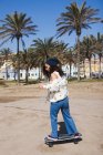 Все тело активной женщины в повседневной одежде катается на скейтборде по дороге вдоль песчаного пляжа и высокие пальмы во время тренировки — стоковое фото